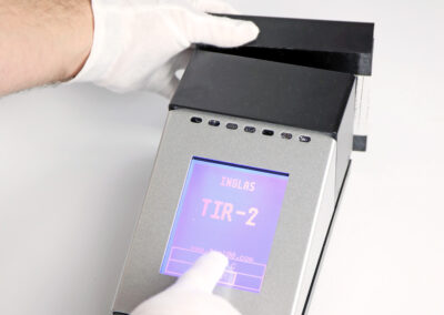 Gloved hand operating the TIR100-2 emissometer touchscreen during an emissivity measurement of a foil sample.