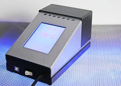 TIR100-2 emissometer with illuminated blue LCD display showing an emissivity reading on a reflective surface.