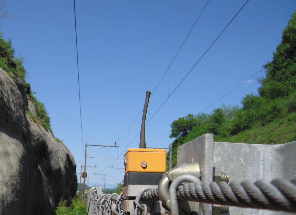 Engineers wearing safety helmets and vests install an Impact Sentinel rockfall monitoring sensor on a protective barrier beside a railway track in a mountainous area.